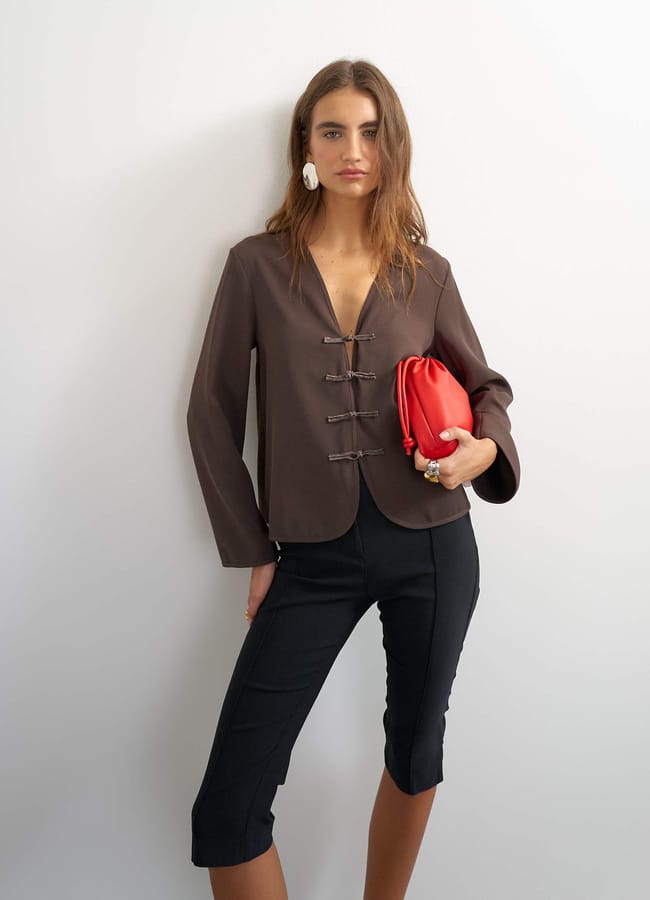 A woman in a brown jacket and black cropped trousers stands against a white wall, holding a red clutch bag and wearing large white earrings—a chic ensemble that perfectly captures the essence of autumn winter 2026 fashion trends.