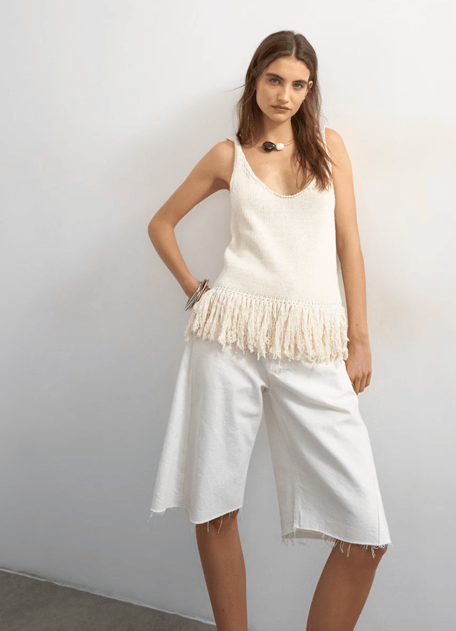 Woman in a sleeveless cream top with fringe detail and wide-leg white culottes stands against a plain white wall, showcasing effortless minimalist summer fashion with her necklace and bracelet, striking a relaxed, casual pose.