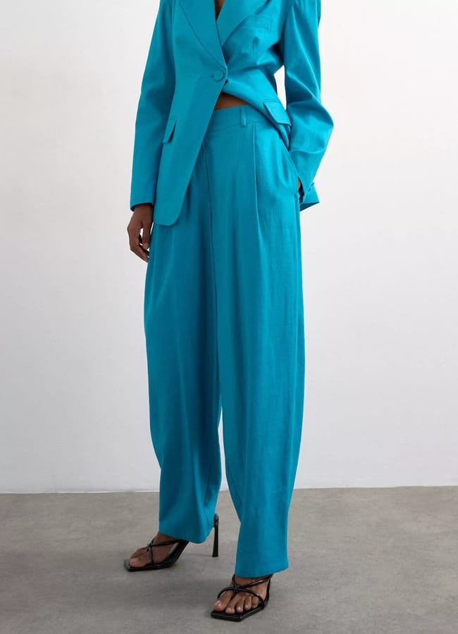 A person wearing a bright blue suit with wide-leg trousers and a blazer exemplifies turquoise fashion, paired with black heeled sandals, standing against a plain light background.