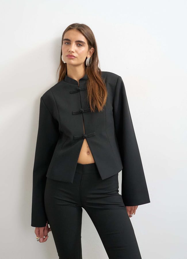 A woman wearing a black, long-sleeve, Mandarin-collar jacket with frog fastenings and matching black trousers stands against a white wall, looking at the camera—a refined look that reflects autumn winter 2026 fashion trends.