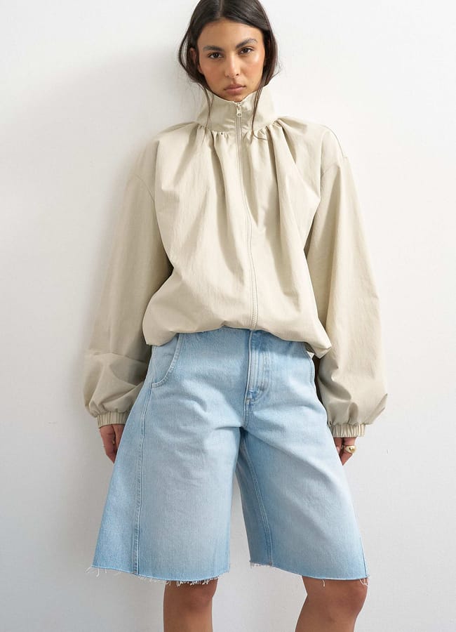 A woman in a beige zip-up jacket with a gathered collar and cuffs, paired with light blue knee-length denim shorts, stands against a plain white wall, looking directly at the camera—an effortless ensemble that nods to autumn winter 2026 fashion trends.