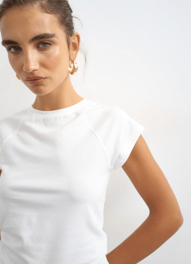 A woman with gold hoop earrings and a white short-sleeve top stands against a plain white background, looking slightly to the side with a neutral expression, her outfit paired effortlessly with classic white denim.