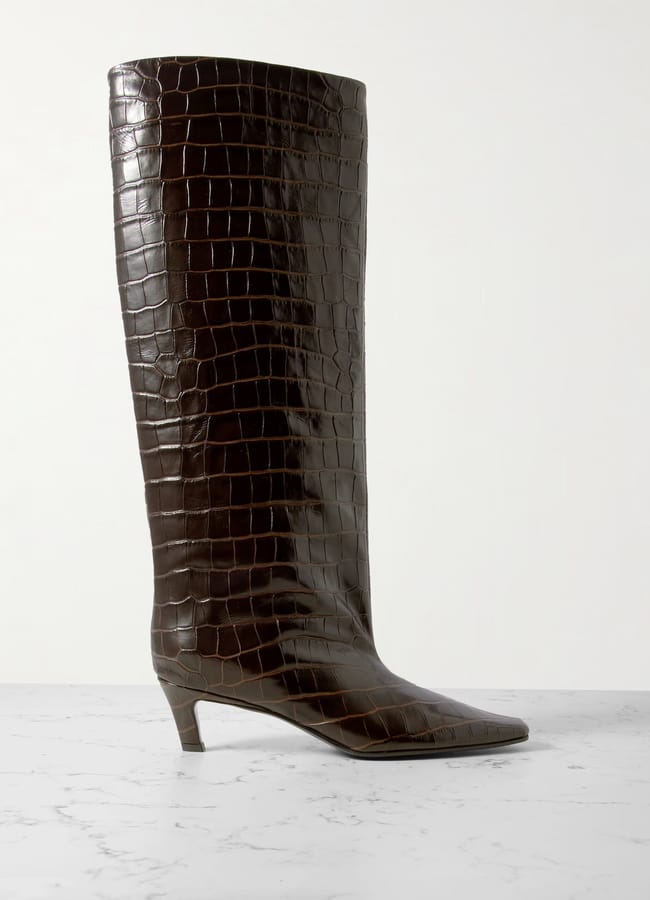 A single dark brown, knee-high boot with a croc-embossed leather texture, a pointed toe, and a low, angled heel is displayed against a light marble surface and white background—perfectly showcased just in time for our Summer Sale.