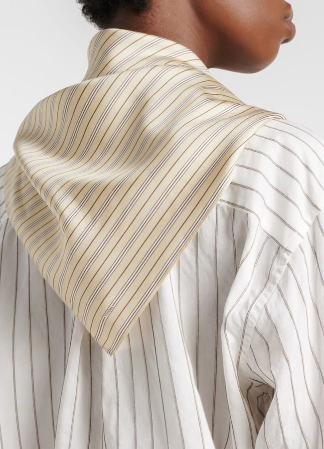 A person seen from behind wears a cream and brown striped scarf over a white shirt with thin vertical stripes. The scarf, perfect for breezy days or as a stylish accent during the summer sale, is draped around the neck with the point hanging down the back.