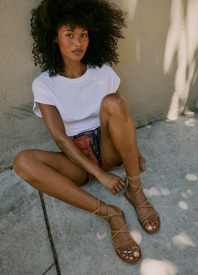 A woman with curly hair sits on a pavement in sunlight, wearing a white T-shirt, red shorts, and tan lace-up sandals, looking at the camera—capturing the carefree spirit of a last minute holiday.
