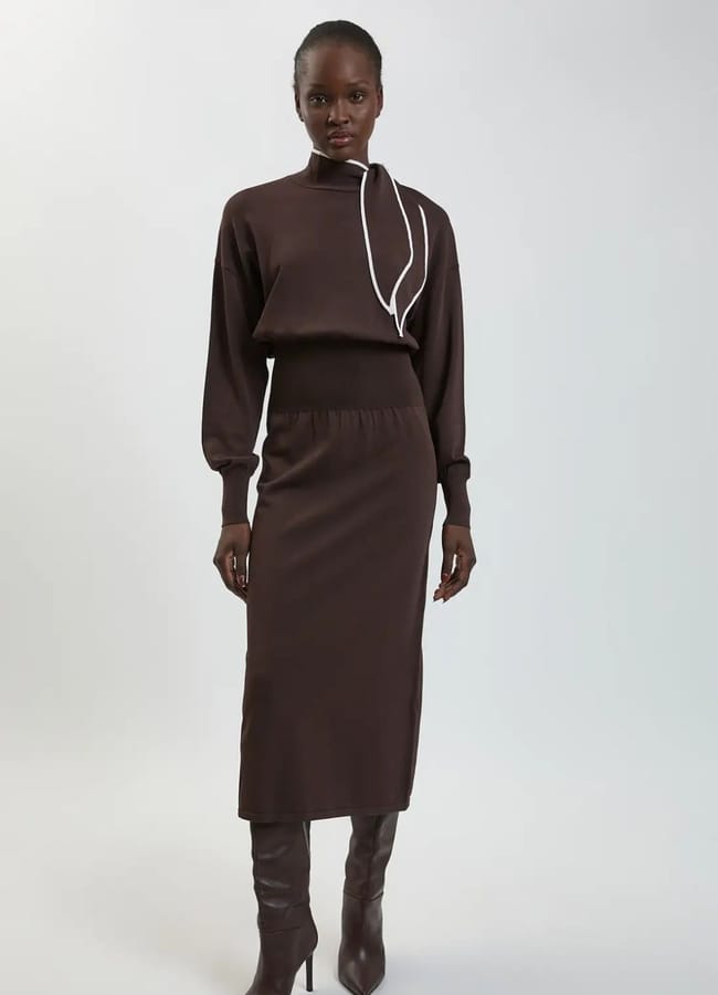 A woman stands in a studio wearing a dark brown long-sleeve top with a white accent and a matching midi skirt, effortlessly capturing the essence of autumn winter 2026 fashion trends. She pairs the outfit with dark brown heeled boots, whilst the background remains plain and light.