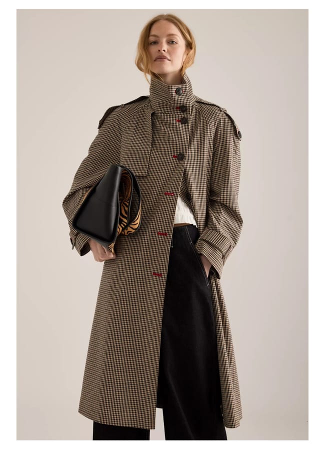 A woman in a long, checked coat with red buttons—perfectly embodying autumn winter 2026 fashion trends—stands holding a large black clutch. She has fair hair pulled back and wears dark trousers and a white top, looking confidently at the camera.