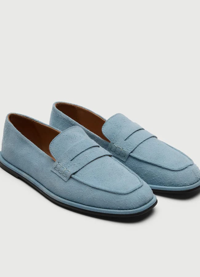 A pair of light blue suede loafers with penny strap detail, black outsoles, and brown interior lining, displayed on a plain white background—perfect for elevating men’s summer dressing with effortless sophistication inspired by The Talented Mr Ripley style.