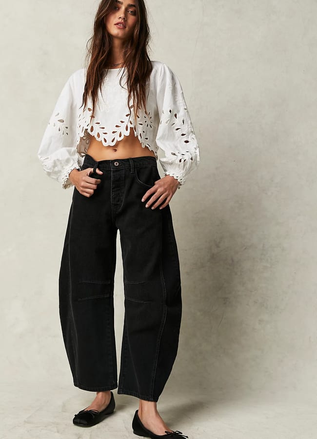 Woman wearing a white, long-sleeve, broderie anglaise crop top and loose black jeans stands against a neutral background, with her hands in her pockets and black ballet flats on her feet—perfect inspiration for those seeking oversized shirt outfit ideas with effortless style.