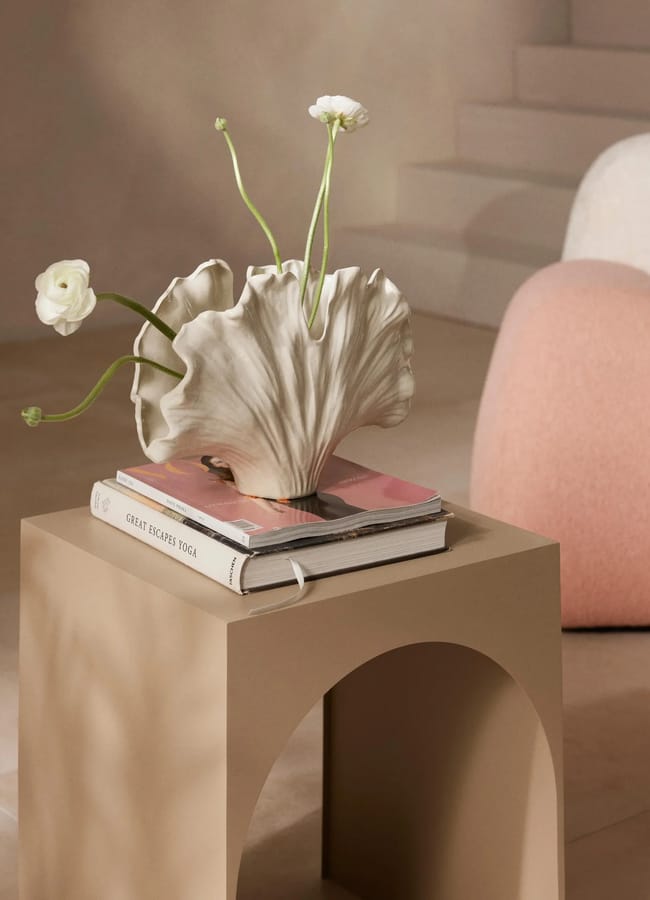 A modern beige side table with a sculptural white statement vase holding three white flowers sits in a softly lit room. Two books lie beneath the vase, and a pink cushioned seat is partially visible in the background.
