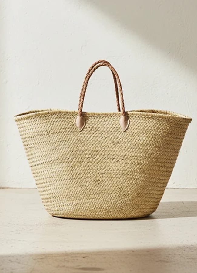 A woven straw tote bag with two plaited handles, sitting on a light-coloured floor against a white textured wall—a perfect accessory for holiday style inspired by celebrity fashion.