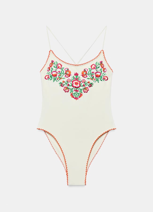 A cream-coloured bodysuit with thin straps features red and green floral embroidery on the chest and scalloped red trim along the neckline, armholes, and leg openings, making it a piece of pretty swimwear perfect for sunny days.