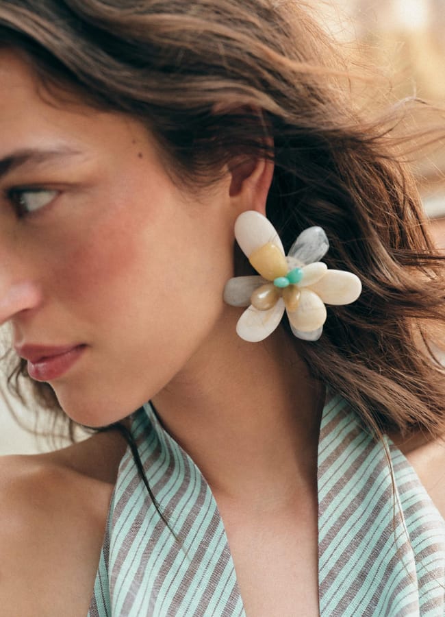 A woman with wavy brown hair wears a large, flower-shaped earring made of petal-like beads in neutral tones and turquoise, paired with a striped halter top—a striking piece of colourful jewellery that perfectly showcases her flair for statement fashion accessories.
