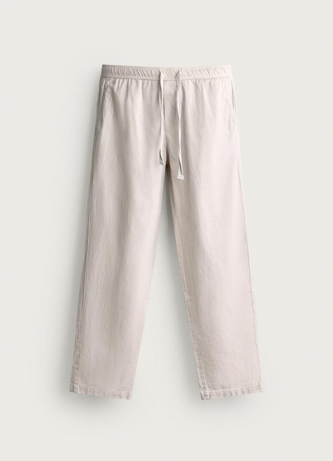 Beige linen trousers with an elasticated waistband and drawstring, displayed flat against a light background. The trousers have a relaxed fit and straight legs, making them a stylish choice for celebrity couples on holiday seeking effortless comfort.