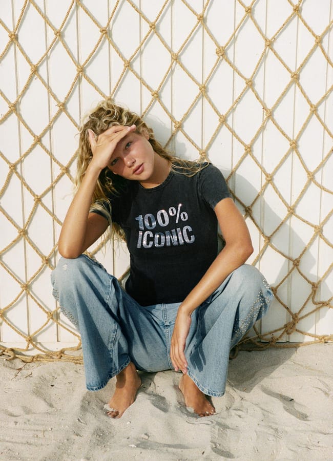 A person sits barefoot on sand in front of a rope net, wearing one of our cool slogan tees—a 100% Iconic T-shirt—and ripped jeans, with one hand on their head.