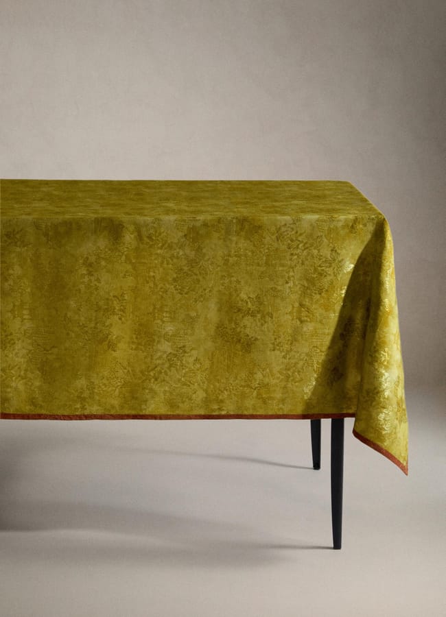 A rectangular table covered with a textured, gold-yellow tablecloth that drapes over the edges, echoing the vibrant chartreuse colour trend. The table has slender, black legs and is set against a plain, light background.