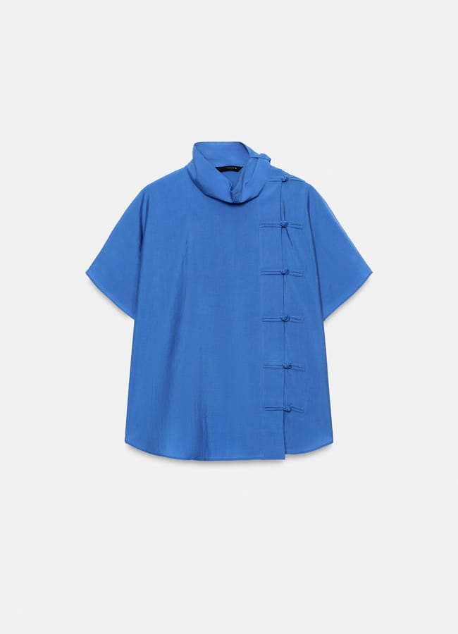 A short-sleeve blue blouse, new in at Zara, features a high collar and five decorative knotted fastenings running diagonally down the front right side.
