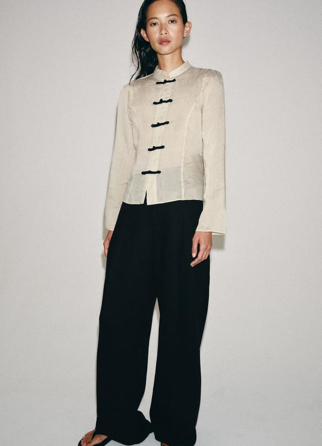 Person standing against a plain wall, wearing a light beige, long-sleeved, mandarin-collar top with black frog fastenings—a style echoing key autumn winter 2026 fashion trends—paired with black wide-leg trousers and sandals. Their hair is down and they have a neutral expression.