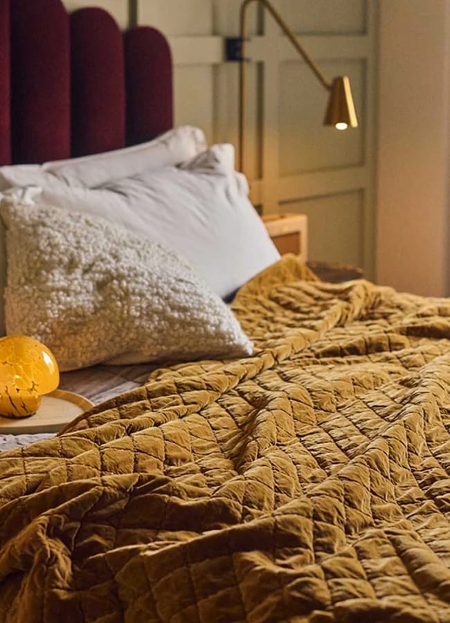 A cosy bed with white pillows, a textured cream cushion, and a mustard yellow quilt. A round yellow lamp sits on a tray, and a brass wall lamp is mounted above the bed in a softly lit bedroom.