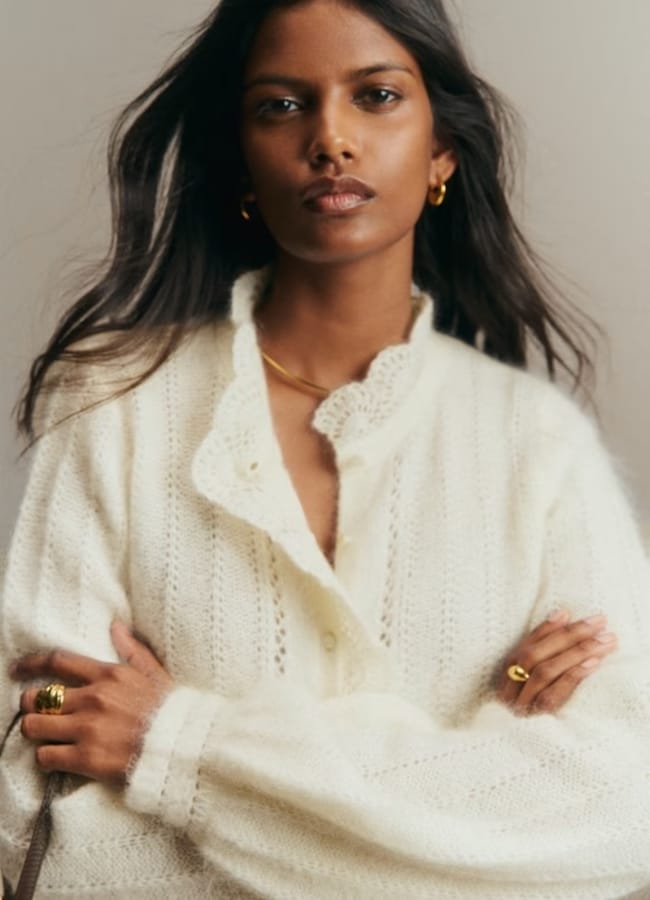 A woman with long dark hair wears a cream knitted cardigan, gold hoop earrings, a gold necklace, and rings, standing with her arms crossed against a plain background—a confident fashion editor known for effortlessly curating chic Paris outfits.