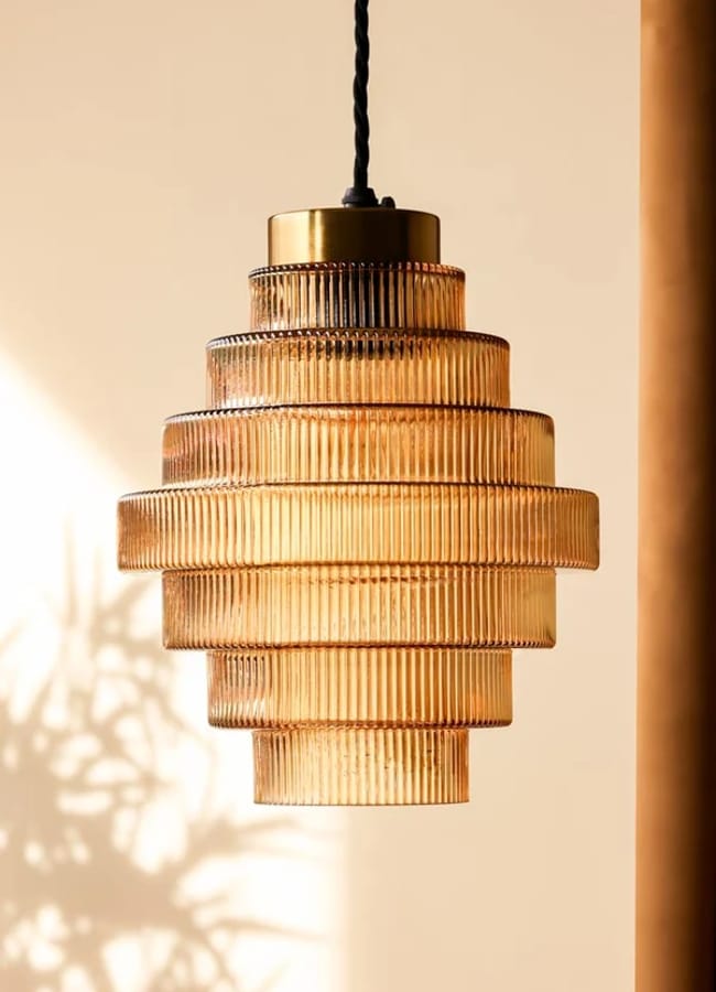 Art deco-style glass pendant light with tiered, ribbed layers hangs from a black twisted flex; warm sunlight casts soft shadows on the beige wall behind it, creating an inviting ambience perfectly suited for any caramel homeware collection.