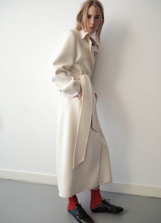 A person stands against a white wall wearing a long cream-coloured coat from one of the top minimalist high street brands, paired with red socks and black shoes, with a neutral expression and hands in the coat’s pockets.