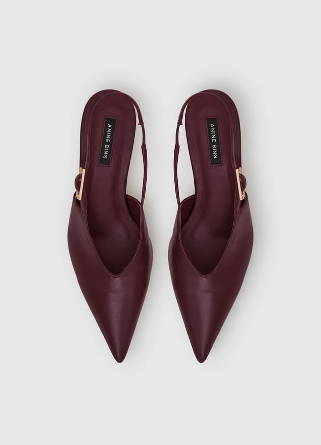 A pair of burgundy pointed-toe slingback flats with gold buckles, reminiscent of sexy heels, viewed from above on a light grey background.