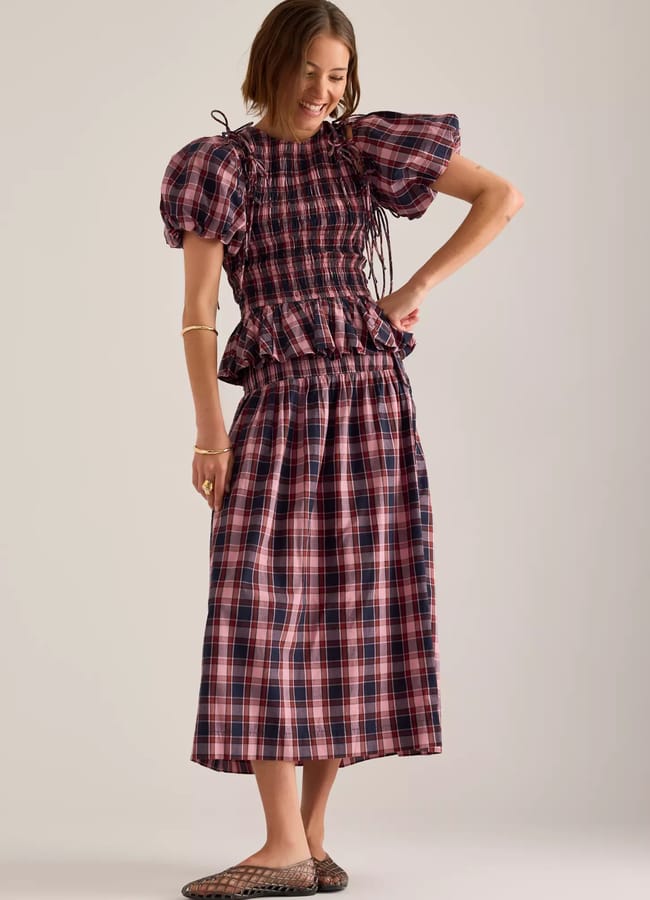 Woman smiling and looking down, wearing a pink and navy checked dress with puff sleeves and a frilled waist, showcasing timeless fashion. She pairs it effortlessly with flat woven shoes, standing against a plain light-coloured background—a perfect choice for those seeking stylish yet comfortable clothes.