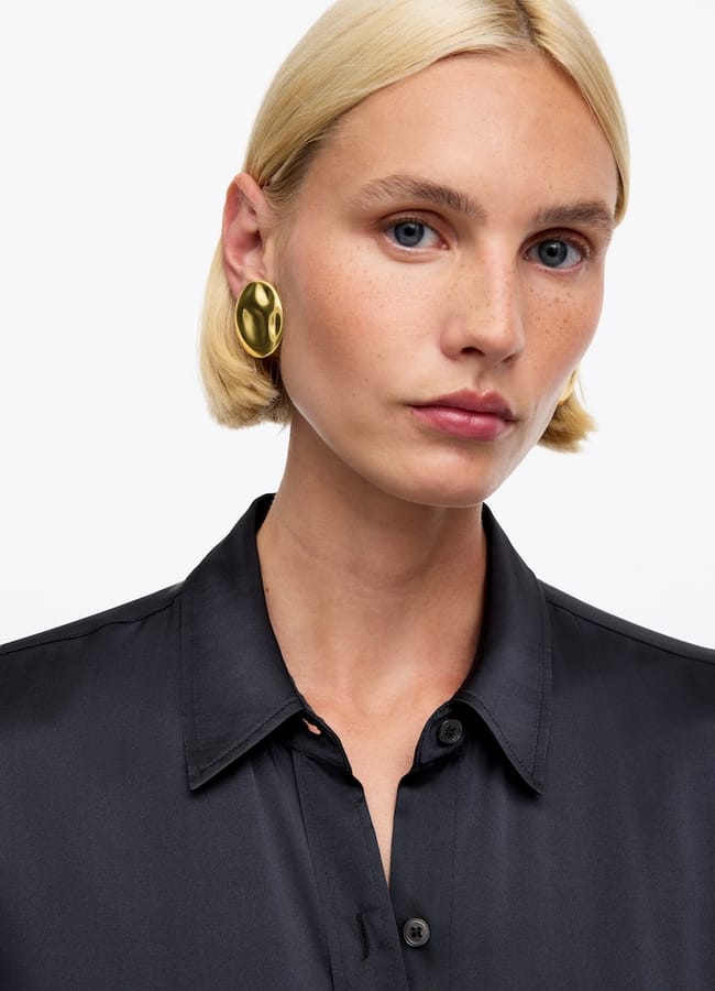 A person with short blonde hair wears a black collared shirt and large gold oval earrings, posing against a plain white background—showcasing how affordable luxury fashion can effortlessly elevate your wardrobe.