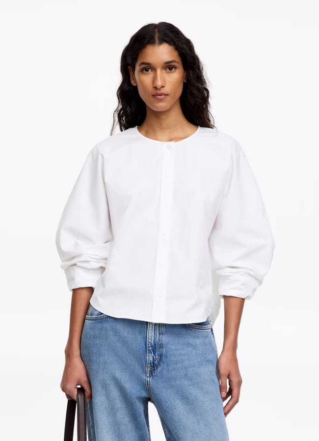 A person wearing a loose white button-up blouse with puffed sleeves and blue jeans, styled in the spirit of minimalist high street brands, holds a black bag while standing against a plain white background.