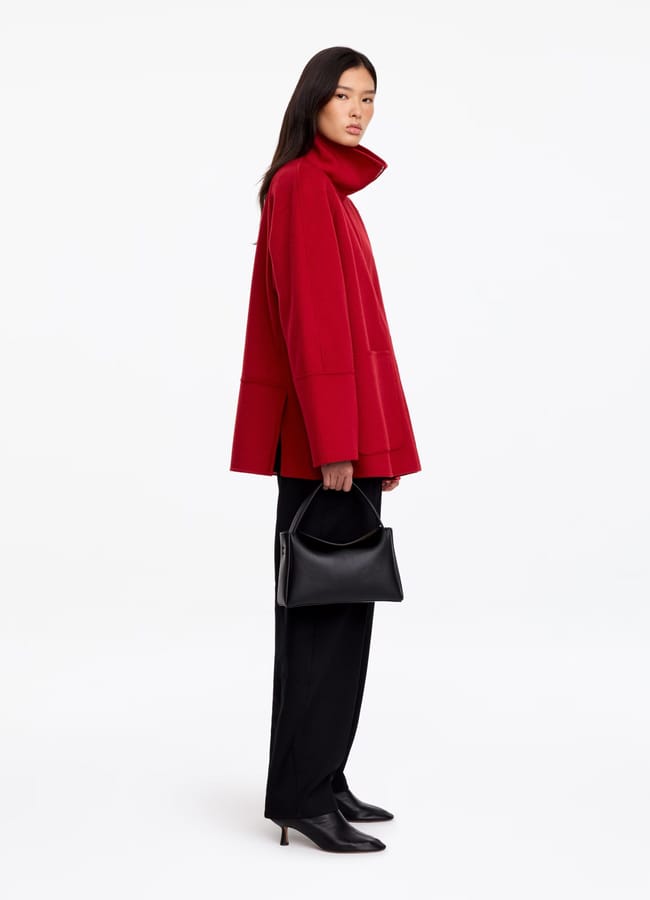 A person stands in profile wearing a stylish red lightweight jacket, black trousers, and black boots, holding a black handbag against a plain white background.