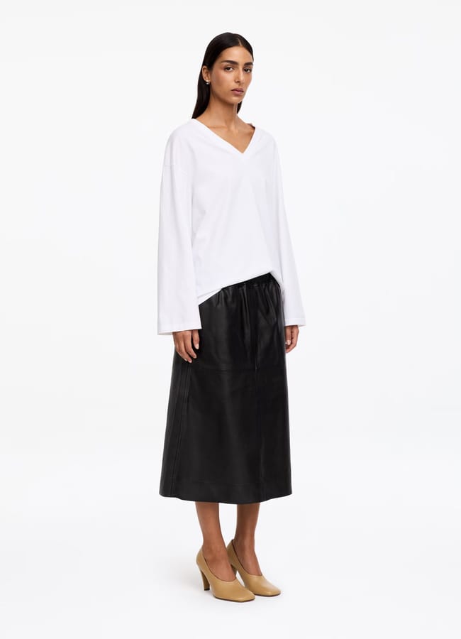 A person stands wearing a loose white long-sleeve V-neck top, a black calf-length skirt, and tan heels against a plain white background, embodying the clean lines and subtle elegance often seen in minimalist high street brands.