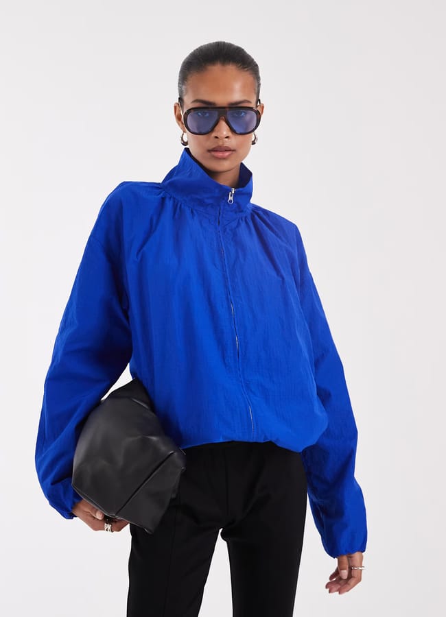 A person wearing a bright blue zip-up jacket reminiscent of International Klein Blue, black trousers, large sunglasses, and silver hoop earrings holds a black clutch bag against a plain light background—a striking pop of colour standing out everywhere in the scene.