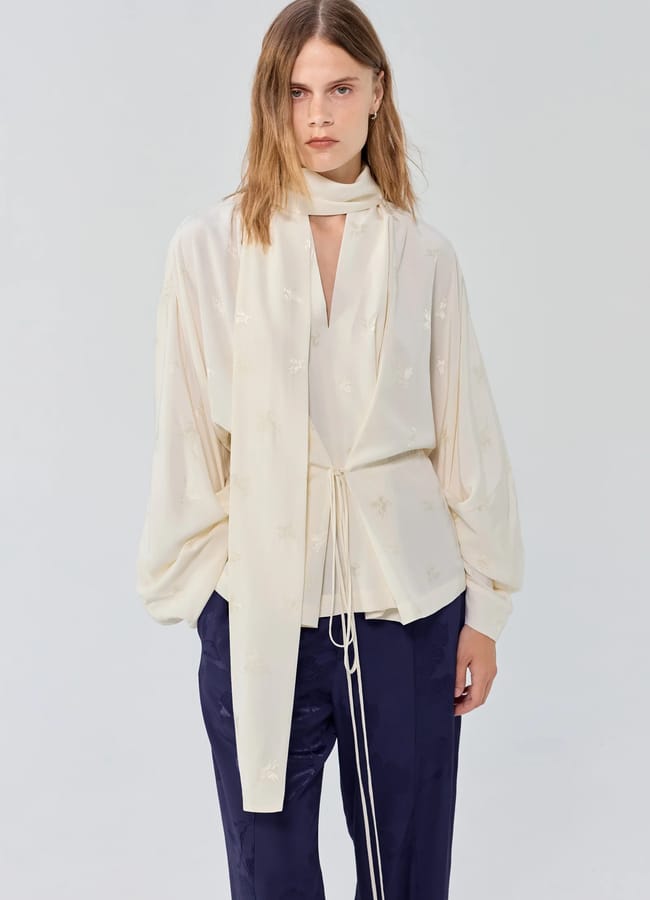 A person with straight, light brown hair wears a cream, long-sleeved blouse with a long tie at the neck and dark blue trousers—an effortlessly chic look that highlights the understated elegance often found in small fashion brands—standing against a plain light background.
