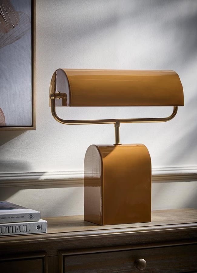 A modern, glossy brown table lamp with a curved shade sits on a wooden surface next to two stacked books, adding a touch of caramel homeware elegance against a light-coloured wall with shadows and part of a framed artwork visible.