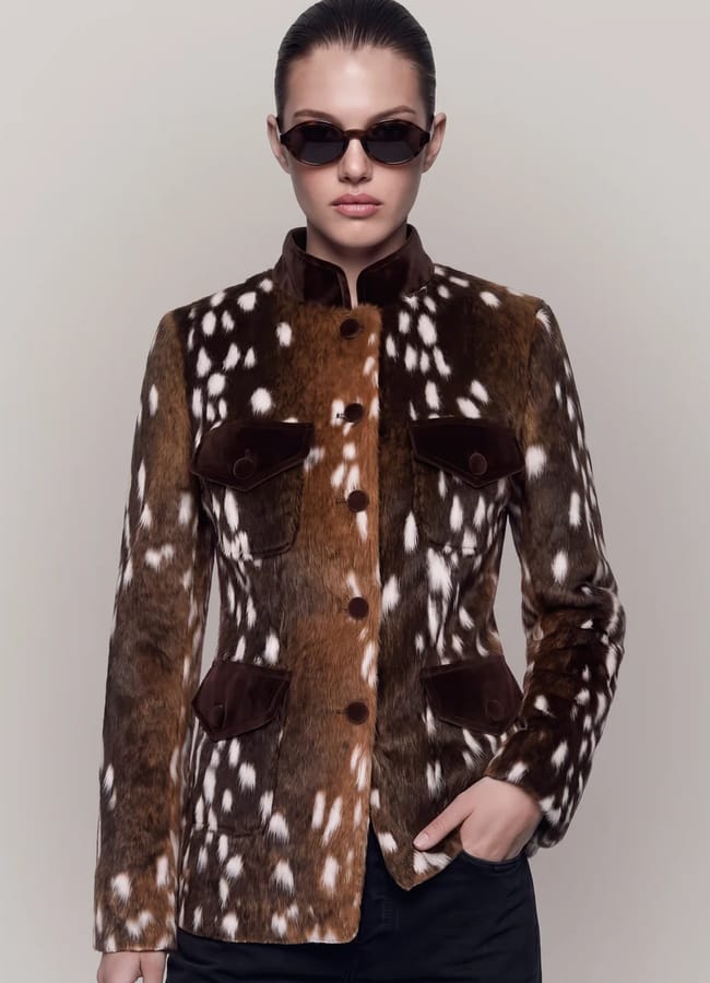 Person wearing a brown and white spotted lightweight fur jacket with button details, black trousers, and dark sunglasses, standing against a neutral background.