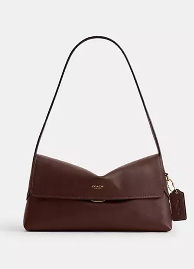 A brown leather shoulder bag with a flap fastening, gold fittings, and a Coach logo on the front. A matching leather tag hangs from the side, making this accessory a perfect choice for those seeking effortless wardrobe upgrades and a touch of expensive fashion.