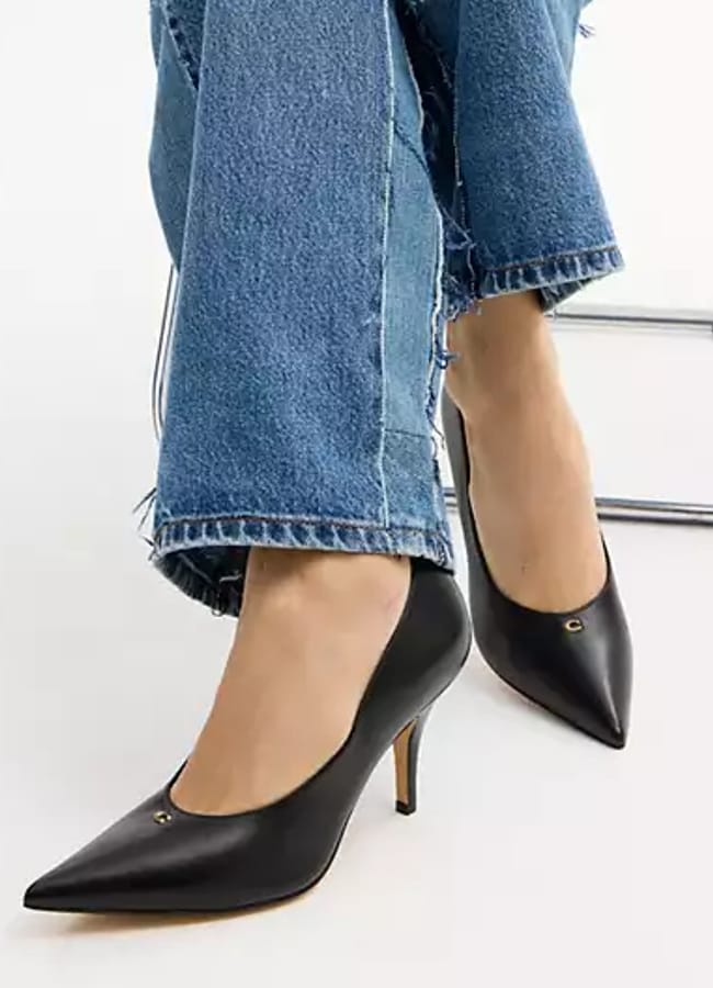 Person wearing frayed hem blue jeans and black sexy heels with pointed toes sits on a chair against a plain background. Only the lower legs and shoes are visible.