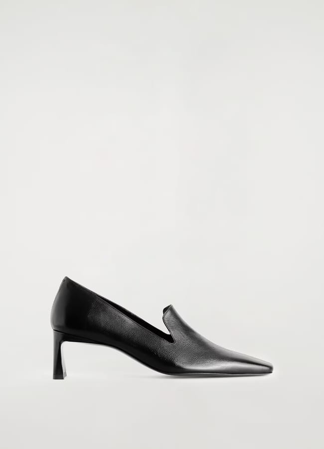 A black leather pointed-toe shoe with a low, angled heel and a subtle V-cut on the upper, shown against a plain light background—a sleek design that echoes the refined aesthetic of minimalist high street brands.