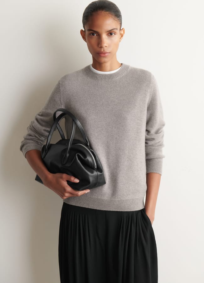 A person stands against a white wall, wearing expensive-looking clothes—a light grey jumper and black skirt—holding a black handbag with one hand and looking directly at the camera, showcasing effortlessly chic wardrobe upgrades and stylish fashion accessories.