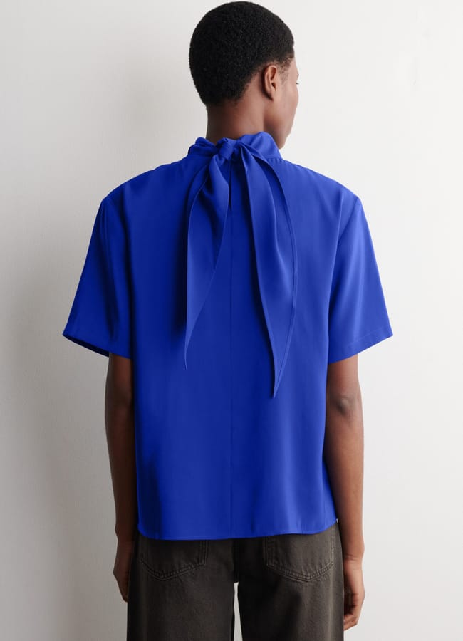 Person standing with their back to the camera, wearing a bright International Klein Blue short-sleeve blouse with a bow tied at the collar and dark trousers, against a plain white background.