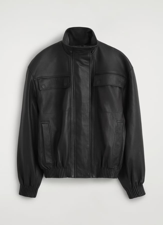 Black leather jacket with a high collar, front zip, two flap chest pockets, two vertical side pockets, and elasticated cuffs and hem—showcasing the refined aesthetic often seen in minimalist high street brands—shown against a plain light background.