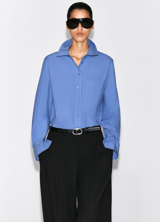 Person wearing a blue collared shirt, black trousers, and black sunglasses stands against a plain light background with hands in pockets, embodying the clean aesthetic often seen in minimalist high street brands.