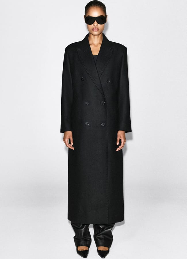 A person wearing a long, double-breasted black coat, black trousers, pointed shoes, and large black sunglasses—an ensemble reminiscent of minimalist high street brands—stands against a plain white background.