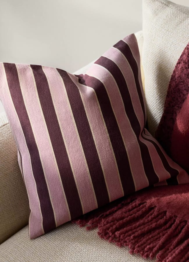 A square cushion with pink and dark burgundy vertical stripes rests on a cream-coloured sofa next to a textured burgundy throw, offering chic homeware under £50 for an effortlessly stylish living space.