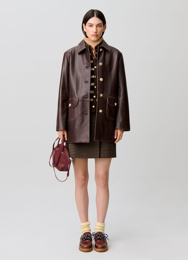 A person wearing a brown leather coat layered over lightweight jackets, paired with a checked skirt and shirt, holding a small maroon handbag, with cream socks and brown shoes, stands against a plain light background.
