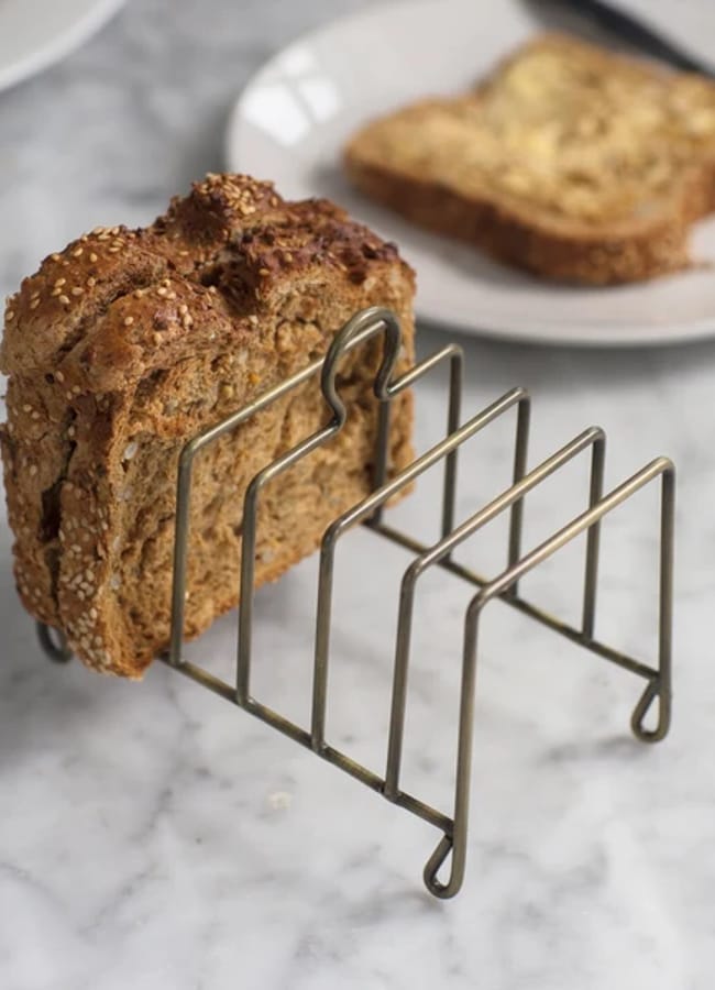 Two slices of wholegrain toast are standing upright in a metal toast rack on a marble worktop, with a plate holding more toast and butter in the background—an inviting breakfast scene elevated by chic homeware under £50.