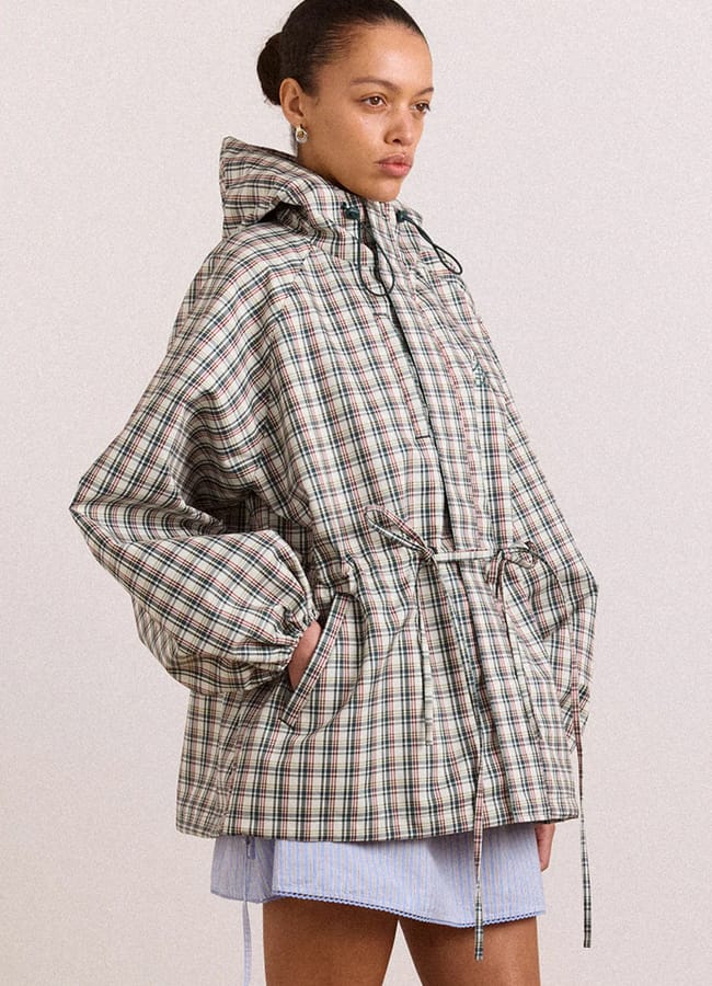 A woman wears a loose, checked hooded jacket with drawstrings and front pockets—one of this season’s essential lightweight jackets—paired with a light blue pleated skirt. She stands against a plain background, looking to the side.
