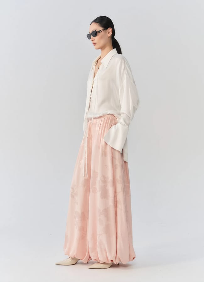 A woman wearing sunglasses, an oversized white shirt, and flowing pale pink floral wide-leg trousers—pieces thoughtfully chosen from small fashion brands—stands against a plain light grey background.