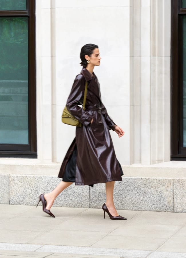 A woman in a shiny dark brown mac and heels walks past a stone building, carrying an olive green shoulder bag—a striking display of fashion that shows how carefully chosen clothes can make any moment memorable.